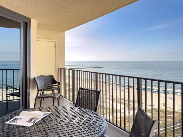 A balcony with a table and chairs, overlooking a beach with umbrellas and a clear view of the ocean.