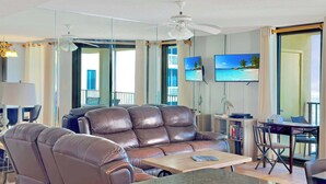 Una sala moderna con un sofá de cuero marrón, una mesa de comedor con sillas, un televisor de pantalla plana en la pared y un balcón con vista al océano.