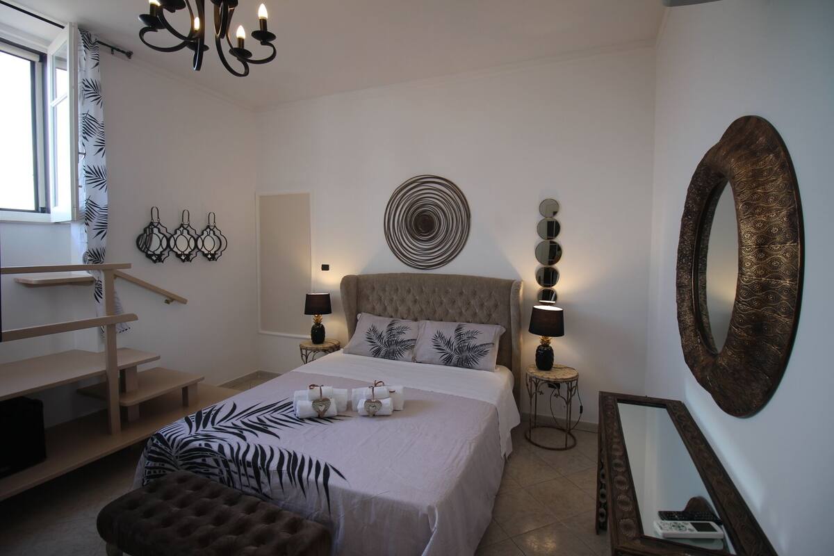 Een slaapkamer met een bed, nachtlampen, een kroonluchter, een grote spiegel en een decoratief wandobject.