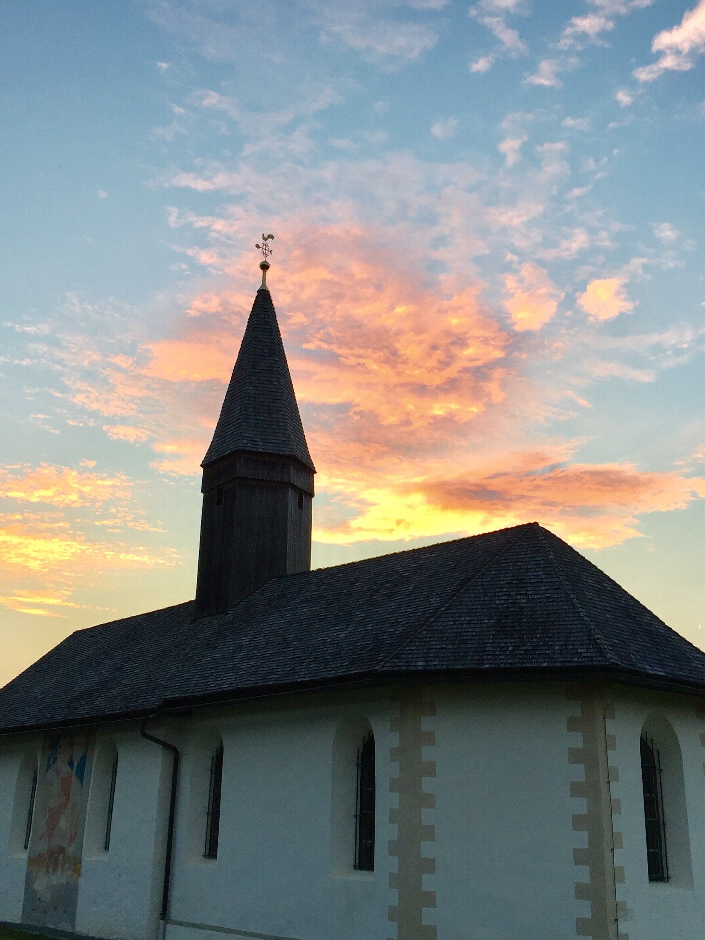 Een kerk met een puntige kerktoren en een kruis bovenop, onder een zonsondergang.