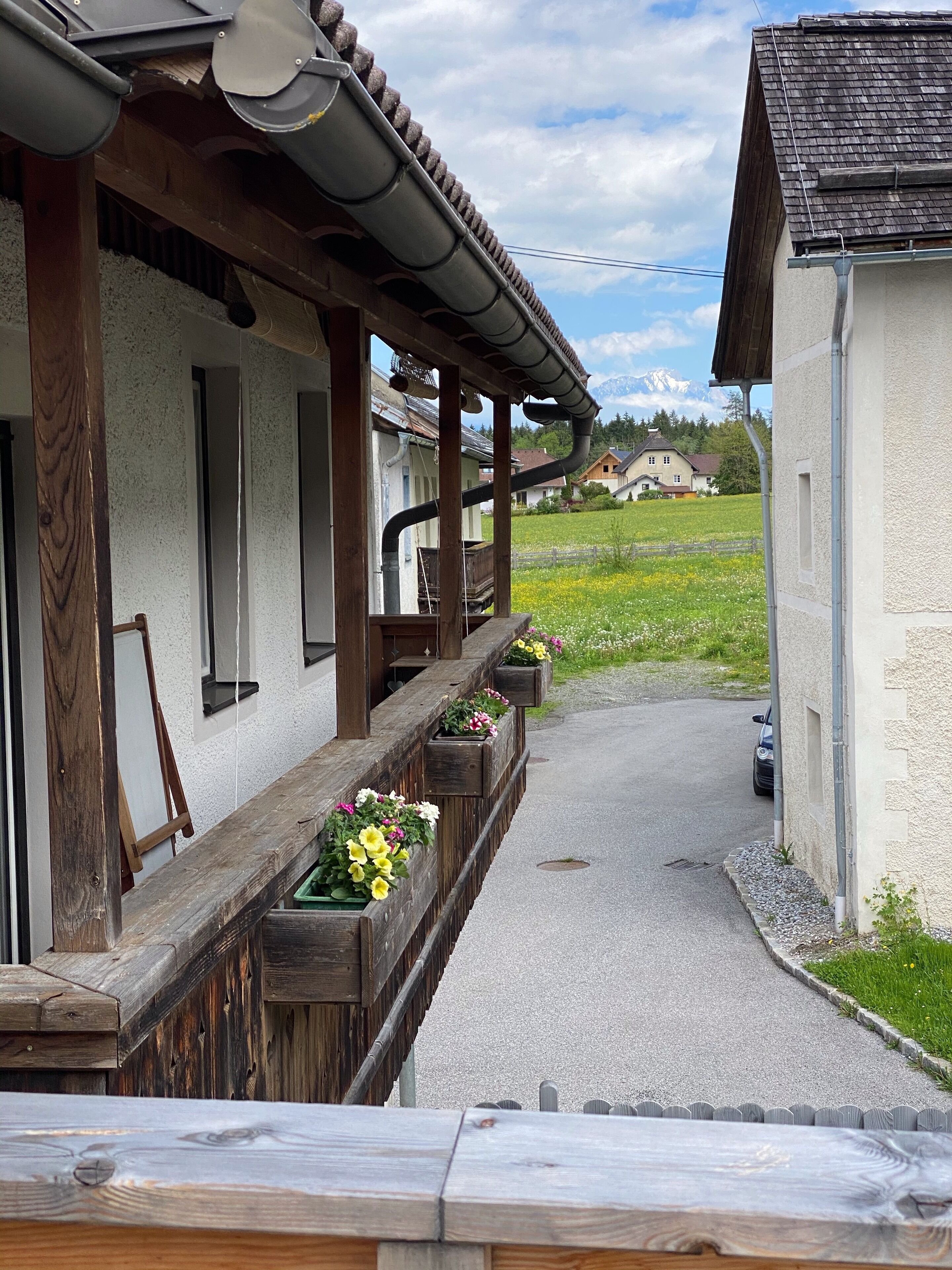 Een gebouw met een houten veranda, bloembakken en uitzicht op een groen veld en bergen op de achtergrond.