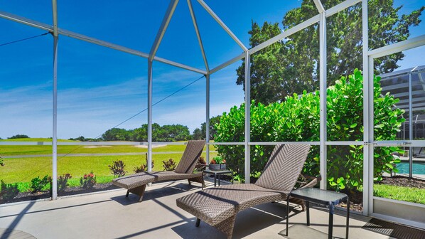 Un espace extérieur abrité, doté de fauteuils de jardin en osier et d’une table, offrant une vue sur une piscine et un paysage verdoyant.