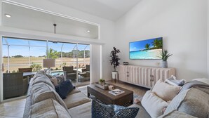 A spacious living room with a large window overlooking a scenic landscape, a comfortable sofa set, a coffee table, and a television displaying a beach scene. - Enjoy Players Club Access in this Luxurious Naples Golf Course Home.   (Naples)