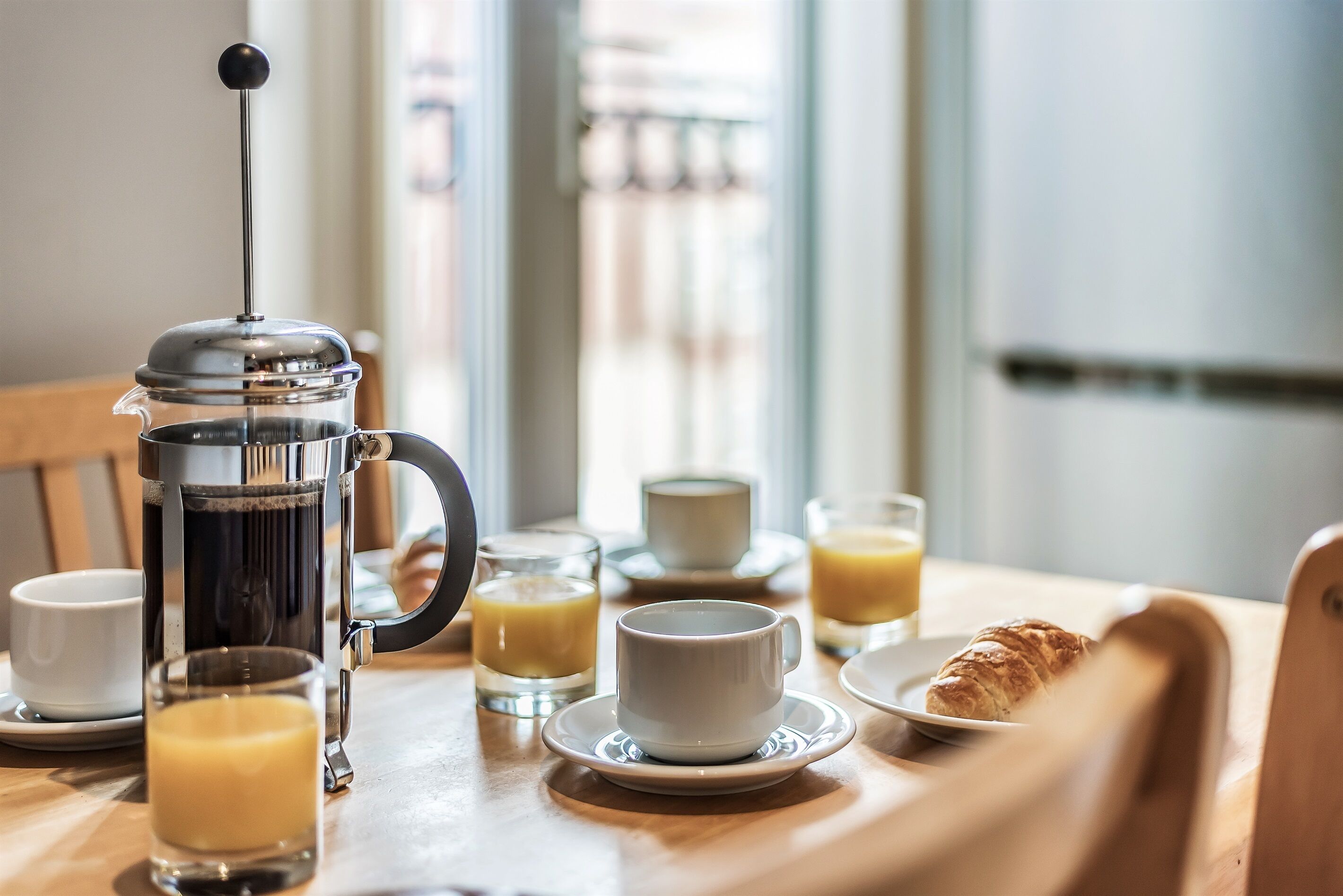 Een ontbijtsetting met een koffiezetapparaat, kopjes, glazen en een croissant op een houten tafel.