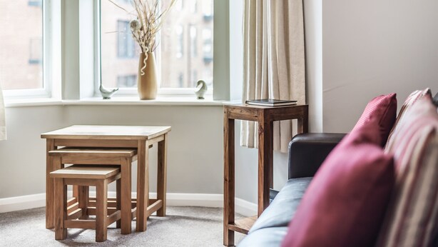 Een gezellige kamer met een raam, een klein houten tafeltje, een bank en een vaas met gedroogde takken.