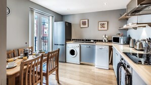 Een moderne keuken met een wasmachine, vaatwasser en magnetron. Er staat een eettafel met stoelen en een klok hangt aan de muur.