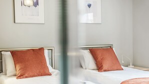 Hotelkamer met twee bedden, elk voorzien van witte beddengoed en een bruin kussen. Er hangen afbeeldingen in lijsten aan de muur, een nachtkastje met een lamp en een raam met uitzicht.