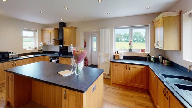 A spacious kitchen with wooden cabinets, a large island, and a view of the outdoors through the windows. - Woodlands Cottage - Rural Retreat - Near York (North Duffield)