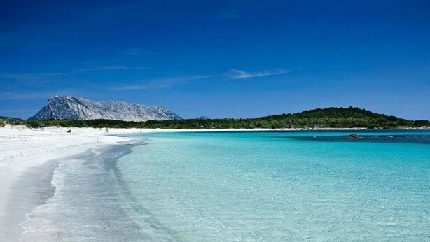 Una playa con aguas turquesas cristalinas, arena blanca y una montaña al fondo.