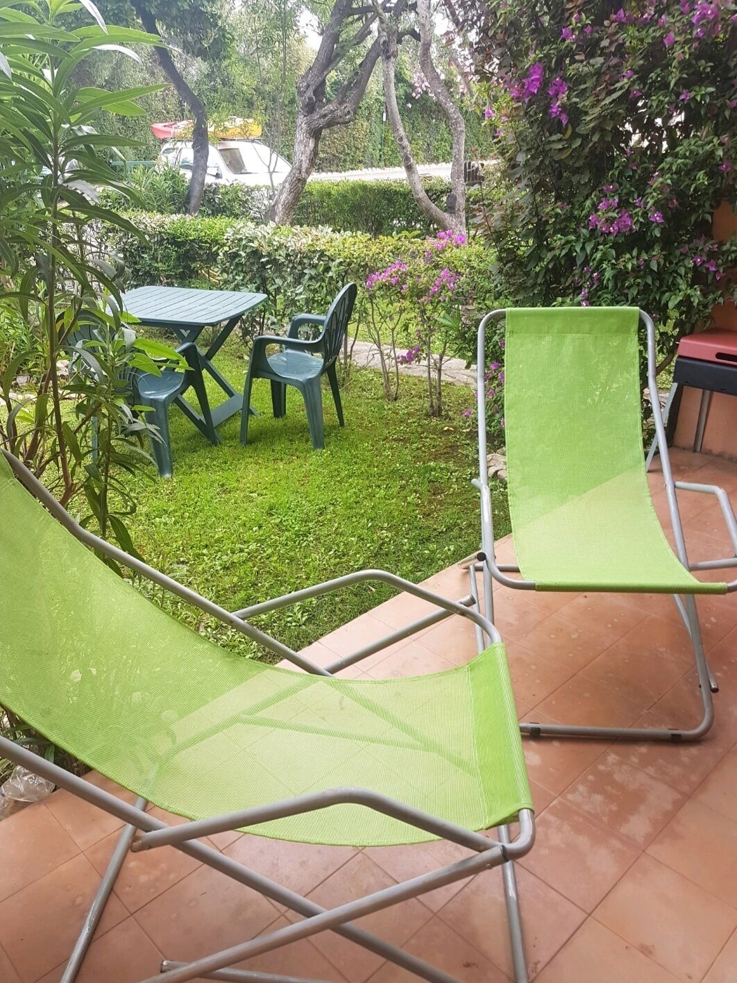 A patio area with green chairs and a table, surrounded by lush greenery and flowering plants.