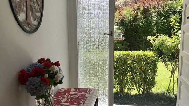 A room with a wooden floor, a red patterned table, a vase with flowers, a decorative wall piece, and a view of a garden through a glass door. - Spacious & gracious (Hamilton)