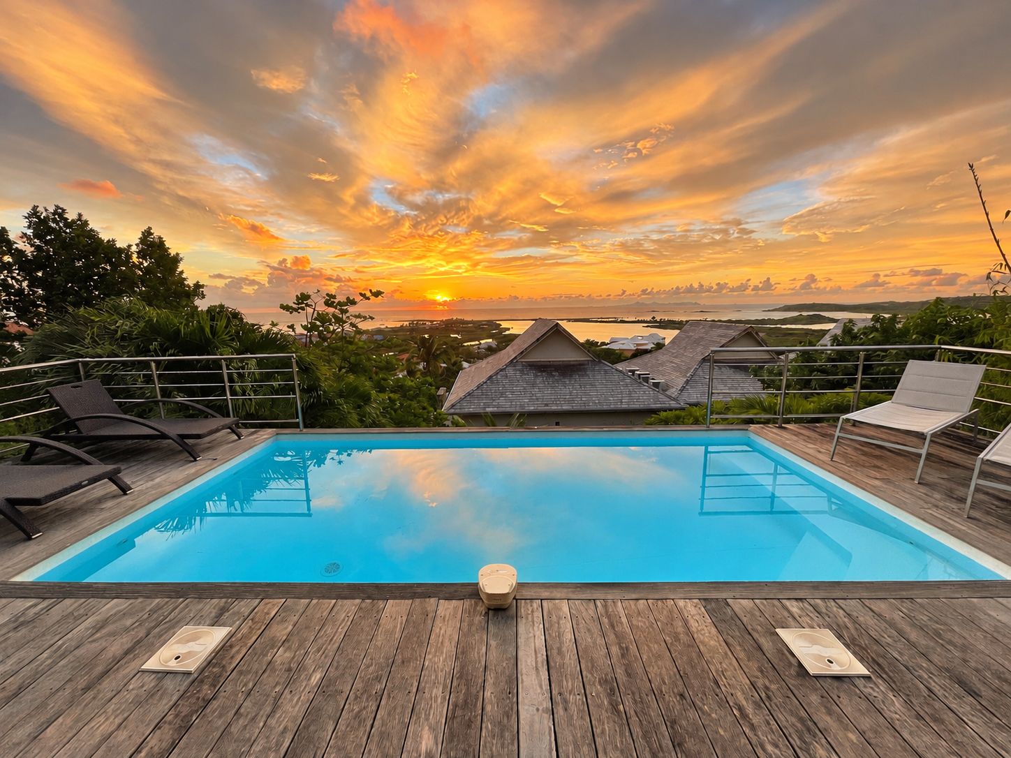 Une piscine dotée d’une terrasse en bois, entourée de chaises longues et offrant une vue sur des maisons et un coucher de soleil.