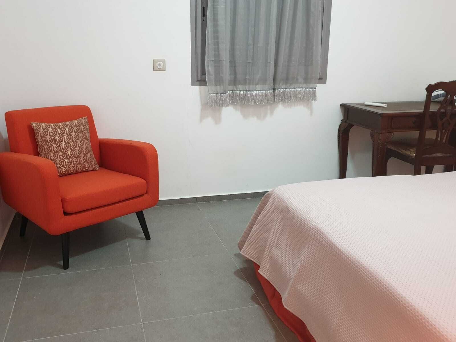 A room with a bed, a red armchair with a patterned pillow, a wooden desk, and a window with a grey curtain.