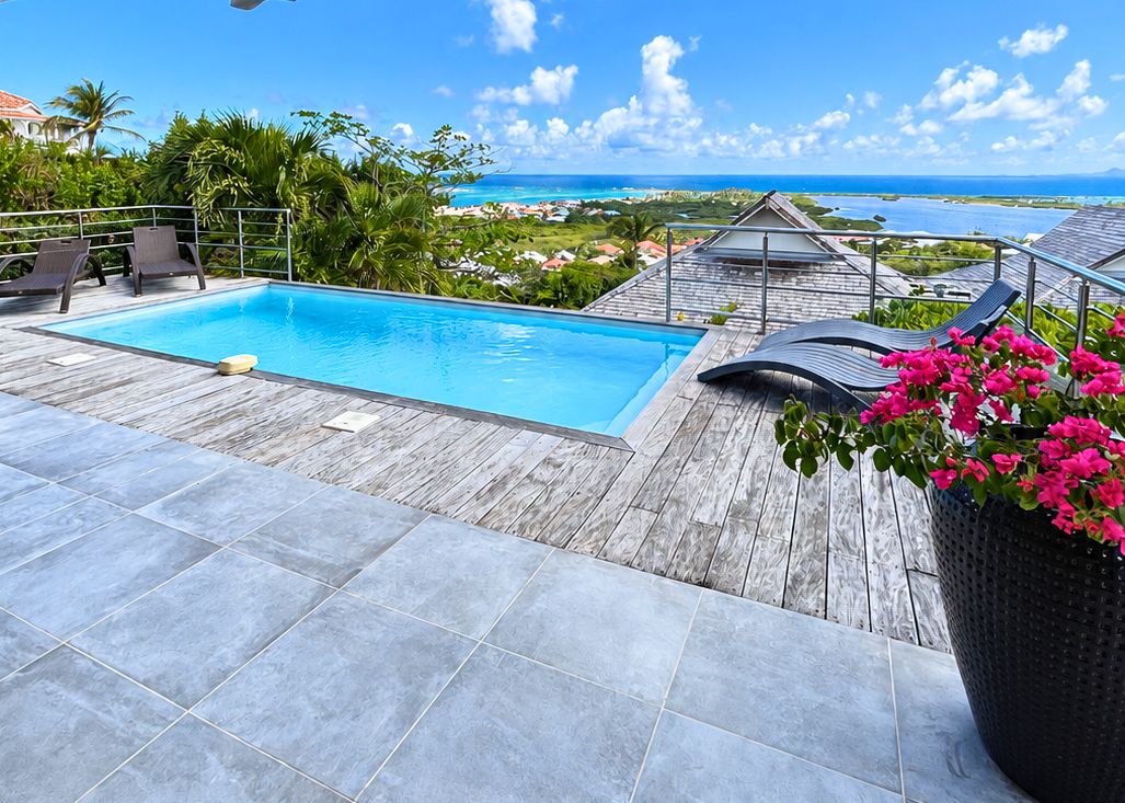 A swimming pool with a wooden deck, surrounded by a railing, and overlooking a coastal landscape.