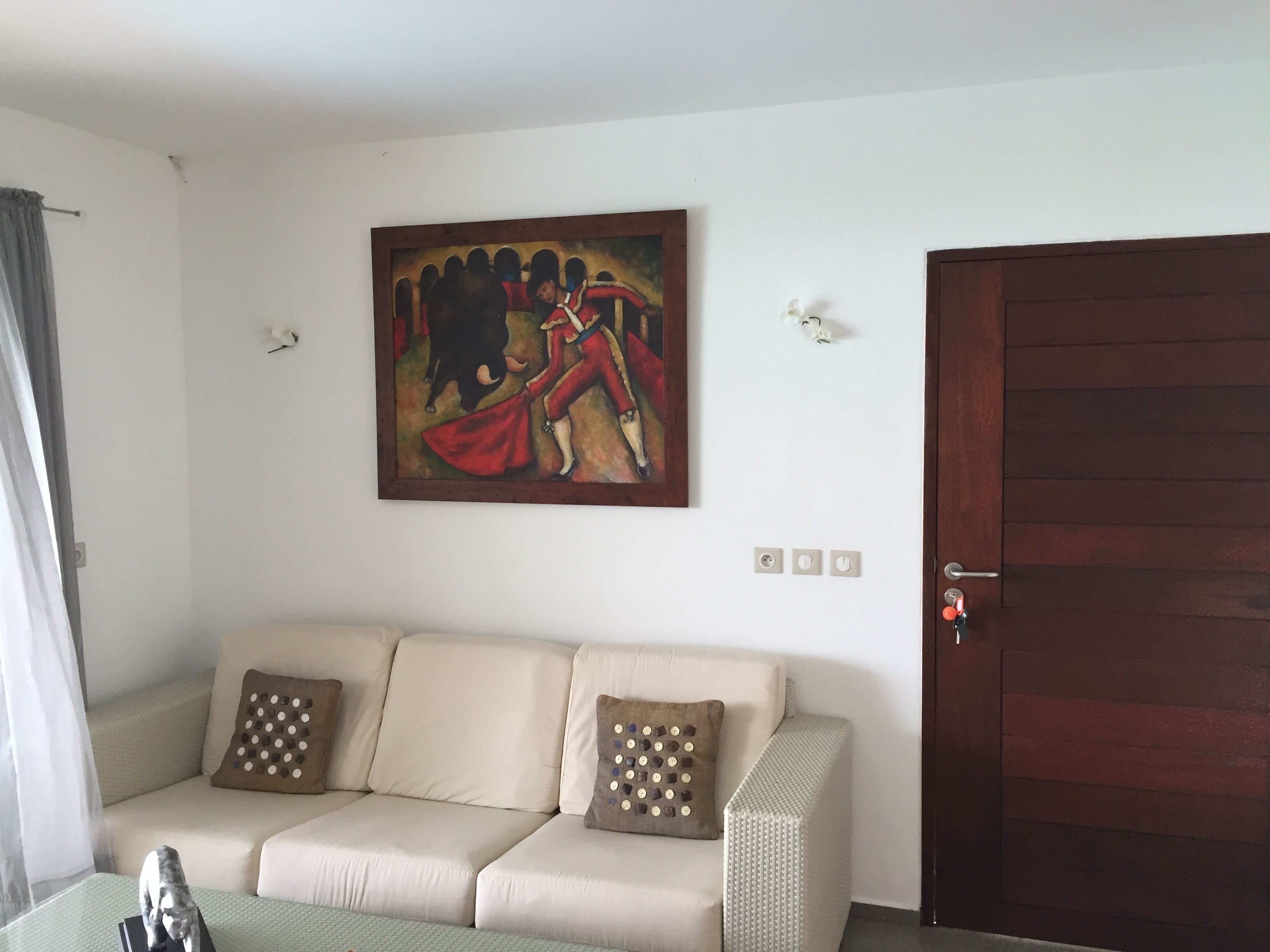 Un salon avec un canapé beige, deux coussins à motifs, un tableau accroché au mur, une porte en bois et une petite table sur laquelle est posée une sculpture.