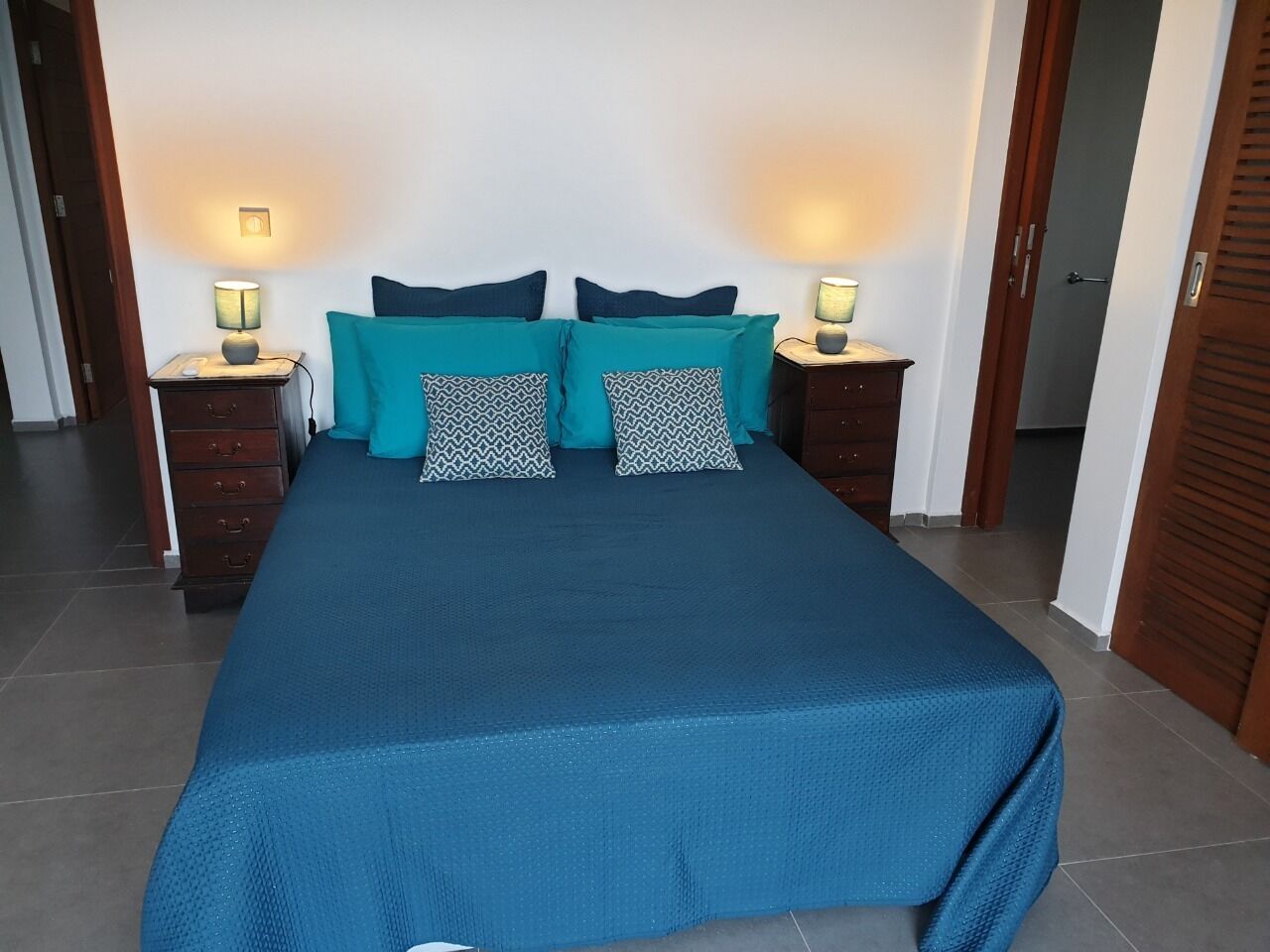 A neatly made bed with a blue bedspread and teal pillows, flanked by two bedside tables with lamps, in a room with tiled flooring and wooden doors.
