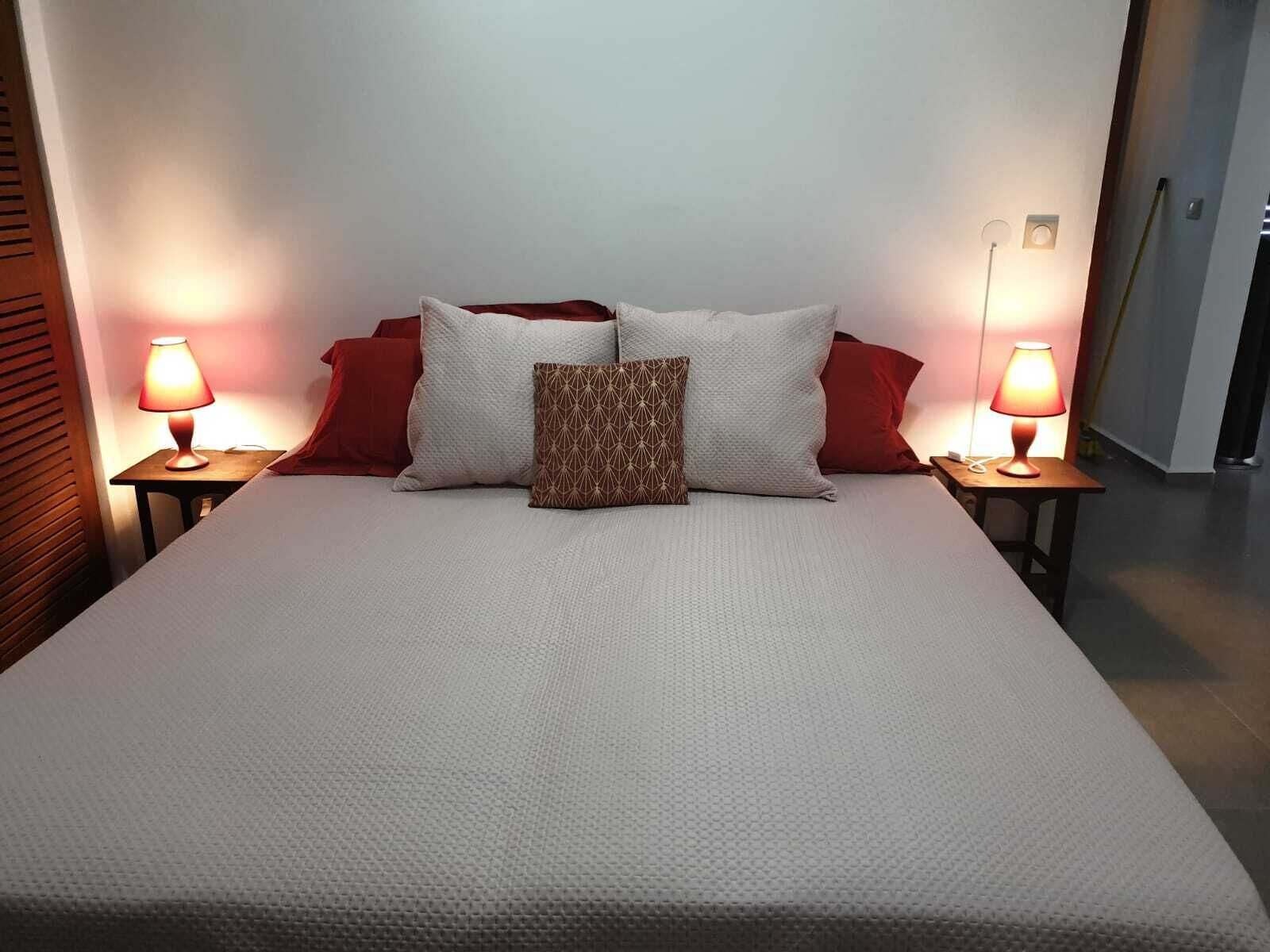 Ein ordentlich bezogenes Bett mit weißer Daunendecke, zwei roten Kissen und einem gemusterten Kissen. Auf den hölzernen Nachttischen stehen jeweils zwei Nachttischlampen.