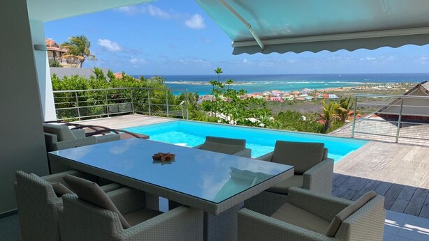 A poolside area with a glass table and wicker chairs, overlooking a beach and ocean view.