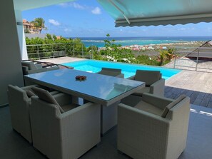 Een zwembadterras met een glazen tafel en rieten stoelen, met uitzicht op een strand en de oceaan.