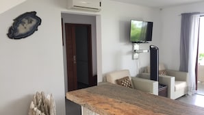 A modern living room with a kitchen area, a flat-screen TV mounted on the wall, and a stone sculpture on the floor.
