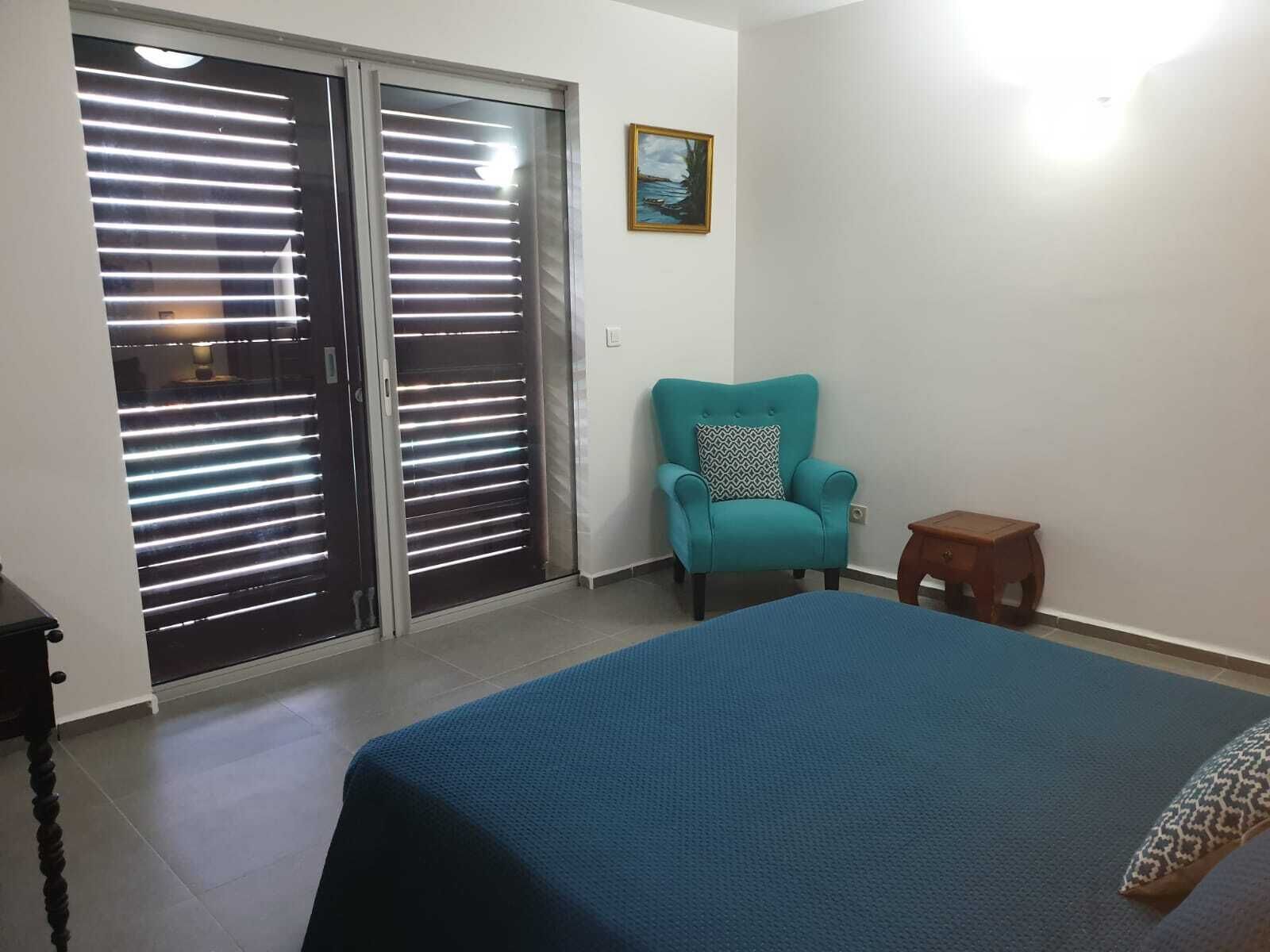 A bedroom with a bed, a chair, a small table, and a sliding glass door with blinds.