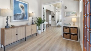 A hallway with a wooden floor, a rattan console table with lamps, a framed painting of a pelican, and a door leading to another room. - Private house with Pool and Spa! (Captiva)