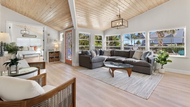 A spacious living room with a high ceiling, a sectional sofa, a coffee table, and large windows offering views of palm trees and a building. - Private house with Pool and Spa! (Captiva)