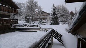 Un balcon enneigé avec une balustrade en bois, une vue sur un immeuble doté d’un balcon, et des arbres couverts de neige.