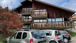 Un bâtiment en bois à deux étages, doté de balcons, avec des voitures garées et un arbuste à fleurs rouges.