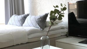 Una cama bien hecha con almohadas grises, un florero de cristal con una rama y un televisor sobre un pedestal.