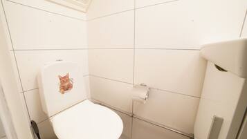 Un baño pequeño con inodoro blanco, un rollo de papel higiénico y una escoba apoyada en la pared.