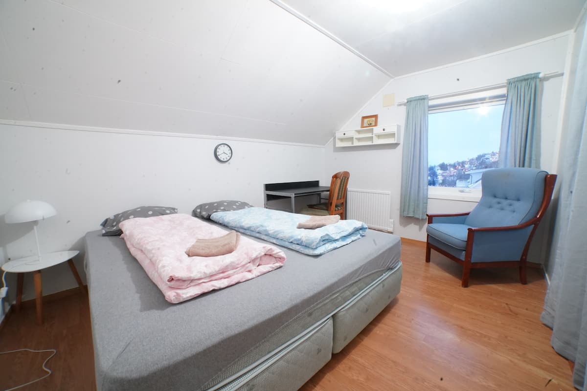 Um quarto com cama, uma cadeira, escrivaninha e janela com vista.