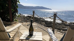 Une table avec une bouteille et des verres, donnant sur une côte rocheuse, avec vue sur l’océan et les montagnes au loin.