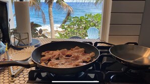 Une cuisine avec une poêle sur la cuisinière, et une vue sur l’océan et les palmiers par la fenêtre.