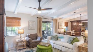 Ein geräumiges Wohnzimmer mit Deckenventilator, einem grünen Hocker, einem weißen Sofa und einem Essbereich mit Holzkohlenstühlen und einem Tisch.