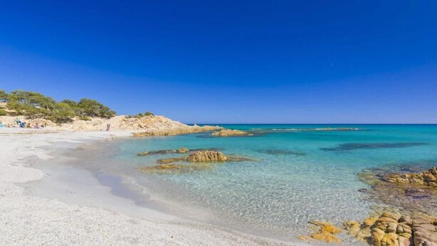 Ein Strand mit klarem türkisblauem Wasser, sandigem Ufer und Felsen.