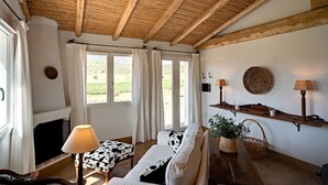 Ein gemütliches Wohnzimmer mit Holzbalkendecke, einer Couch, einer Lampe, einem Kamin und Blick ins Freie.