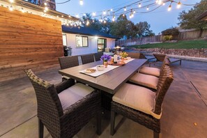 Un patio al aire libre con una mesa de madera y sillas de mimbre, luces colgantes y una pared de piedra al fondo.