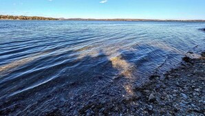Un lac aux eaux limpides, bordé d’une rive rocheuse et entouré d’arbres qui se dessinent à l’horizon sous un ciel bleu.