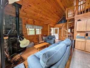 A log cabin interior with a stone fireplace, wooden furniture, a staircase, and a kitchen area. - Secluded Log Cabin by Saltwater Cove - Wildlife Haven (Sullivan)