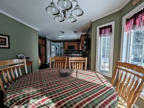 A room with a plaid bedspread, wooden chairs, a dining table, a kitchen area, and a window with curtains. - Woodland House w/ Deck, Near Beaches & Acadia (Hancock)