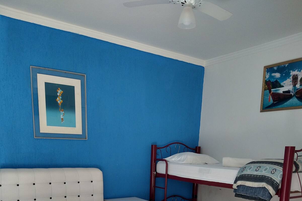 Une chambre d’hôtel avec un lit simple, un lit superposé, un petit bureau et un ventilateur de plafond.