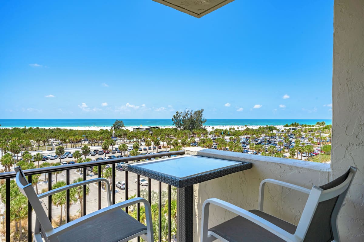 Un balcon avec une table et des chaises donnant sur une plage et un parking.
