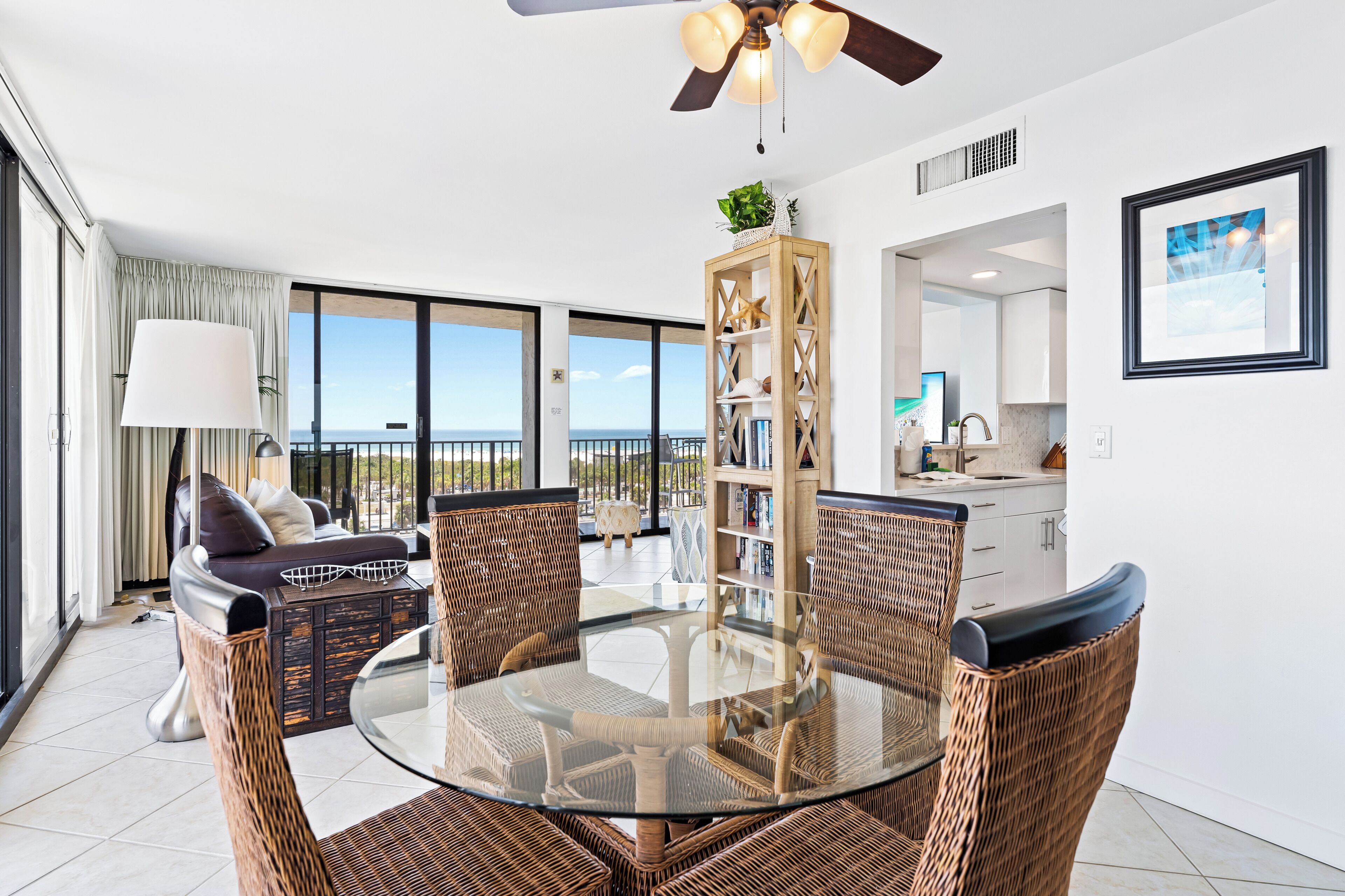 Uma sala de estar moderna com uma mesa de jantar de vidro rodeada por cadeiras de vime, um sofá e uma grande janela com vista para o mar.