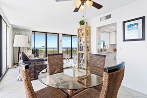 Un salon moderne avec une table à manger au plateau en verre, entourée de chaises en osier, d’un canapé et d’une grande fenêtre offrant une vue sur la mer.