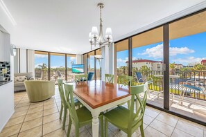 A dining area with a wooden table and green chairs, large windows with a view of a pool and buildings, and a chandelier. - Our House at the Beach by Beachside Mgt (Siesta Key)