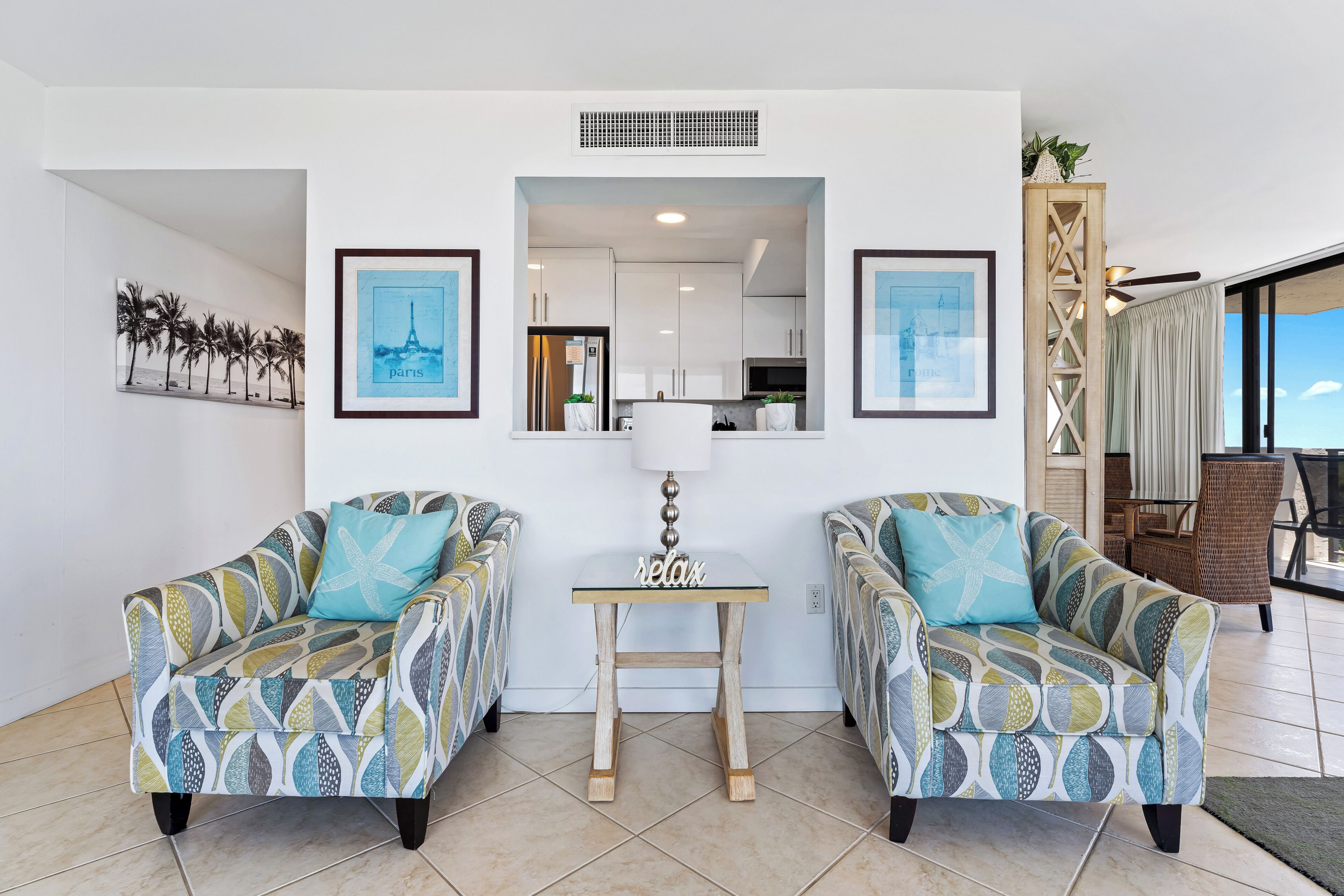 A living room with two patterned armchairs, a small table, and a view of the kitchen through an open doorway.