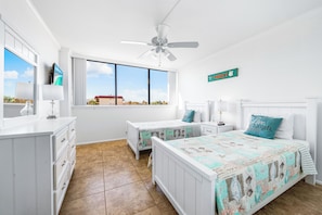 A bedroom with two beds, a ceiling fan, a large window with a view, and a sign on the wall. - Our House at the Beach by Beachside Mgt (Siesta Key)