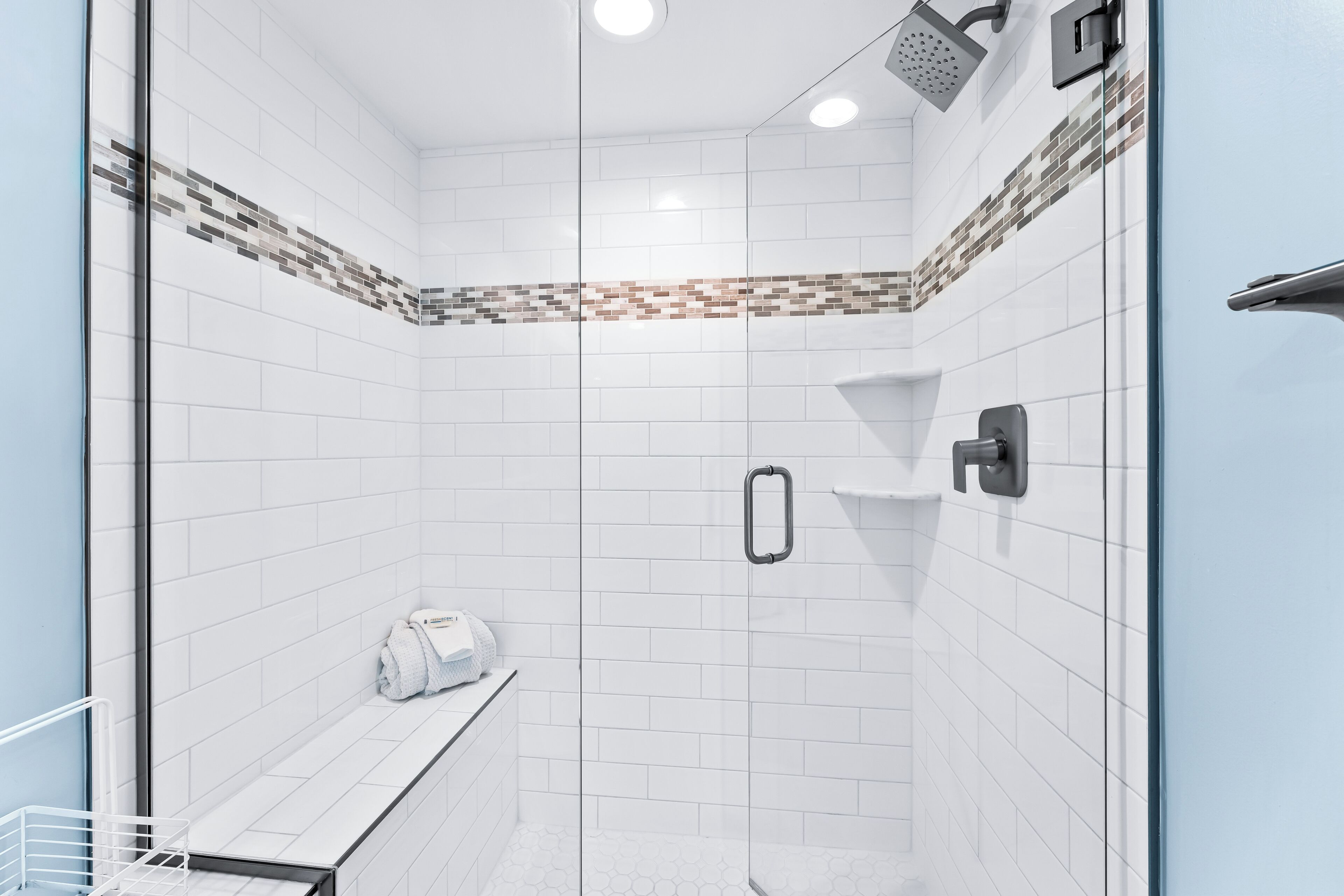A modern shower with a glass door, white tiles, and a small bench with towels.