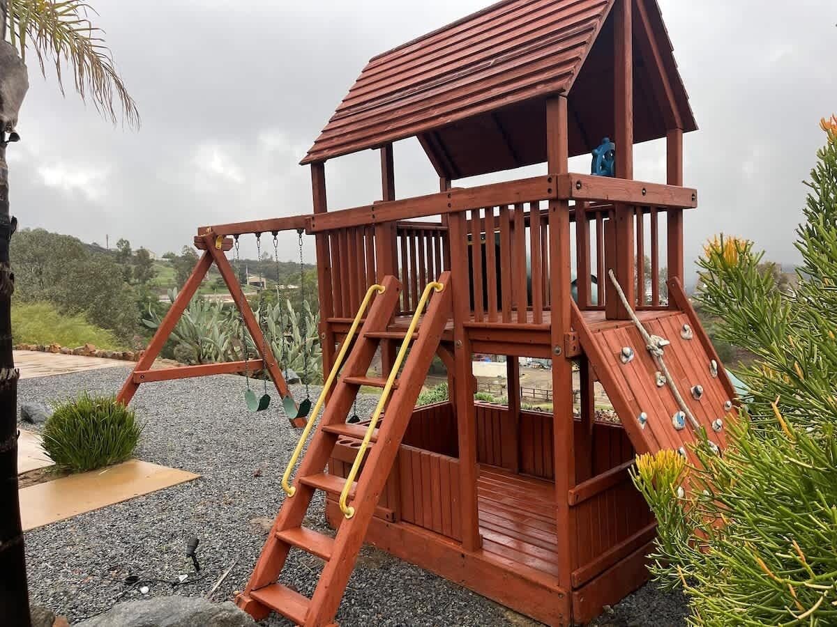 A wooden playhouse with a slide and ladder, surrounded by gravel and greenery.